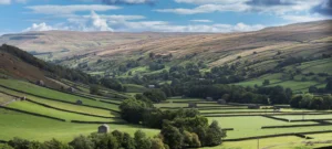 Rolling green hills with dry stone walls, scattered stone barns, and grazing sheep under clouds.