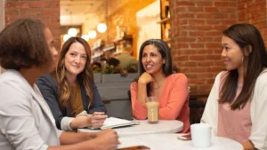 A diverse group of women sitting around a table, talking and supporting each other in a cafe setting.