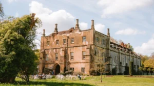 A historic stone manor house with large windows and tall chimneys set against a green lawn.