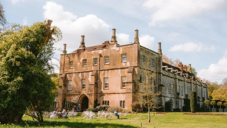 A historic stone manor house with large windows and tall chimneys set against a green lawn.