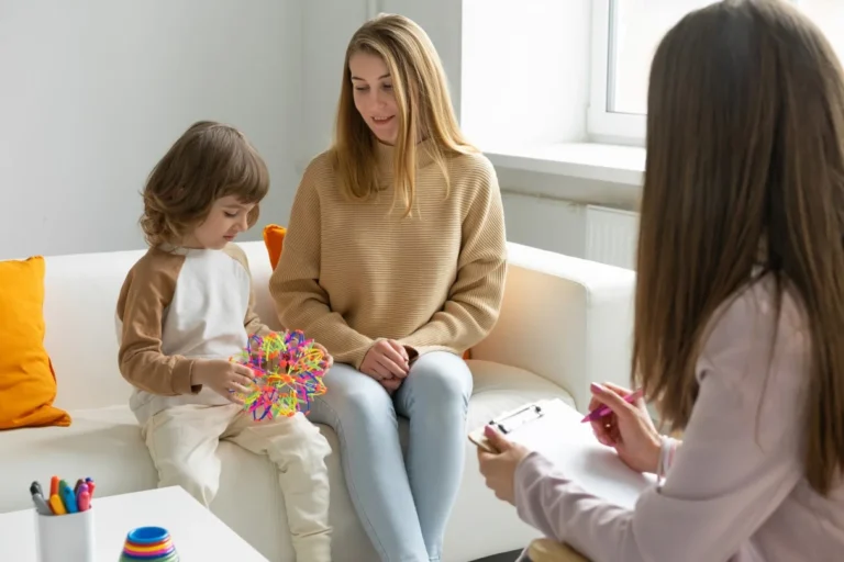 A mother and child sit on a sofa while a therapist takes notes during an appointment.
