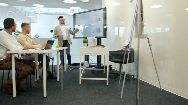 A business professional presenting data and charts on a large monitor to colleagues in a modern office setting.
