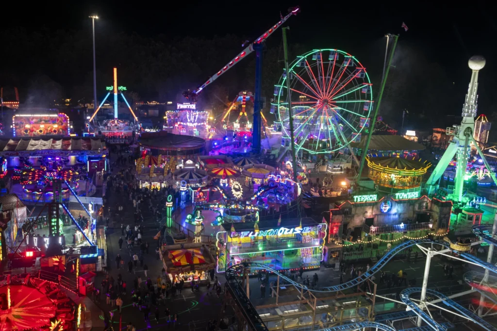 Busy night fairground with colorful neon lights, crowds of people, and a lit-up Ferris wheel.