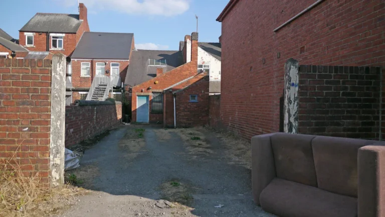 A discarded brown sofa sits in a residential alleyway between brick houses.