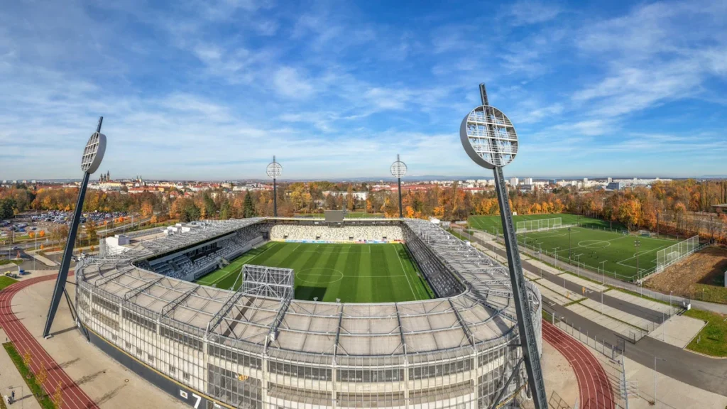 A panoramic aerial view of a large sports stadium and surrounding training pitches under a blue sky.