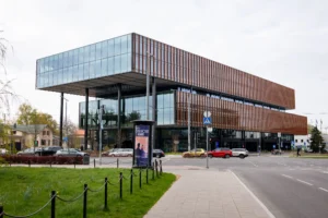 Modern glass office building with a large cantilevered upper floor overlooking an urban street intersection.