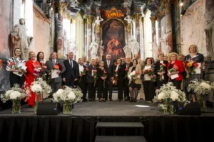People holding bouquets and awards stand on a stage inside a grand, baroque-style church hall.