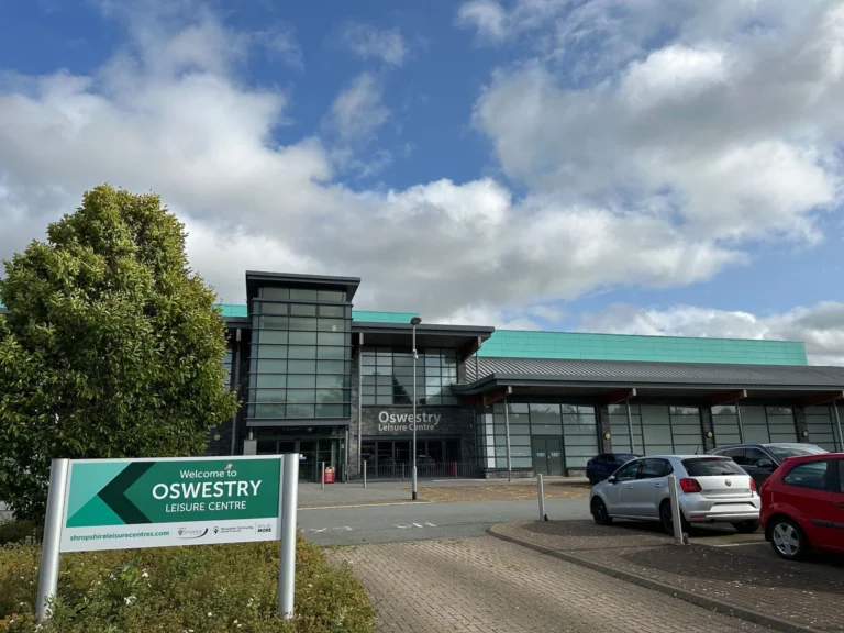 Oswestry Leisure Centre building behind a green sign with parked cars under a blue, cloudy sky.