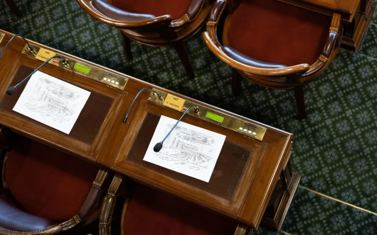 Overhead view of wooden legislative desks with microphones, documents, and ornate red upholstered chairs.
