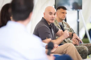 A bald man holding a microphone speaks during a panel with a soldier in camouflage.