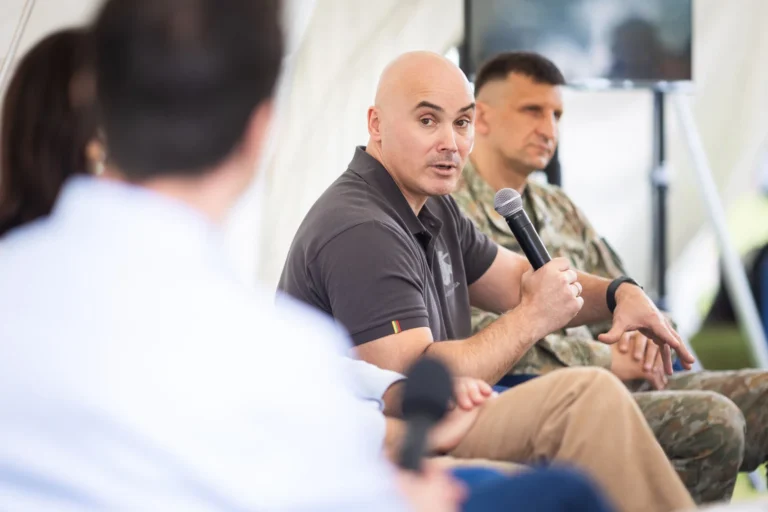A bald man holding a microphone speaks during a panel with a soldier in camouflage.