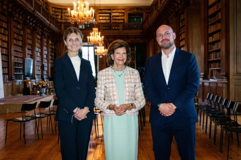 Queen Silvia with two officials in a historic library featuring tall bookshelves and ornate chandeliers.