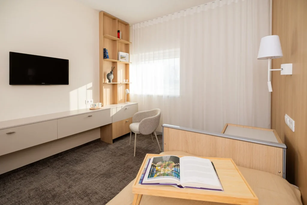 Modern interior featuring light wood furniture, a wall-mounted TV, a desk, and a bed with a book.