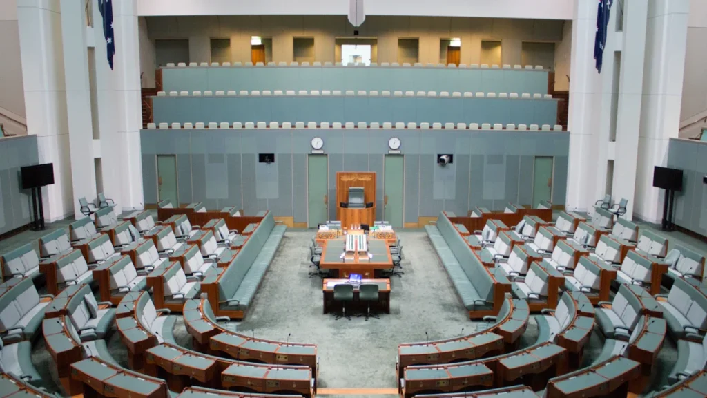 A large government chamber with green benches and wooden desks in a British parliamentary layout.