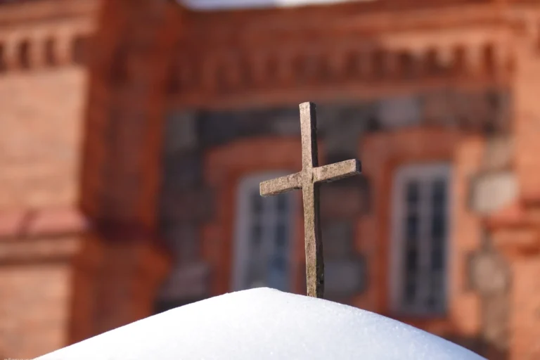 Small stone cross standing in fresh snow with a blurred red brick building in the background.