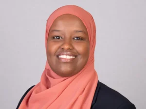 Smiling woman with dark skin wearing a peach hijab and black blazer against grey background.