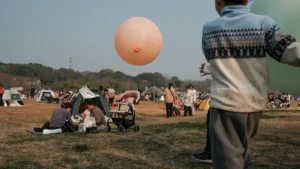 A child playing with a large balloon at a crowded outdoor park event with families and tents.