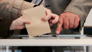 A close-up photograph of a person casting a ballot into a clear voting box while an official points toward the slot.
