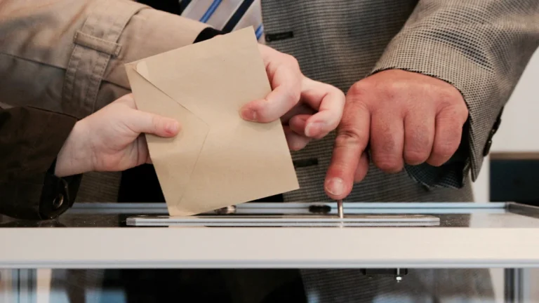 A close-up photograph of a person casting a ballot into a clear voting box while an official points toward the slot.
