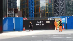 Construction workers in safety gear stand outside a gated entrance at a metropolitan building site.