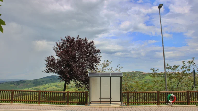 A glass bus stop shelter stands on a roadside overlooking vast, rolling green hills under clouds.