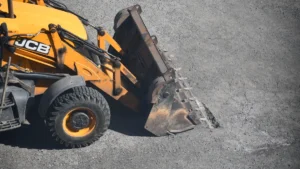 A yellow JCB backhoe loader digging up a damaged road surface as part of scheduled road repairs.