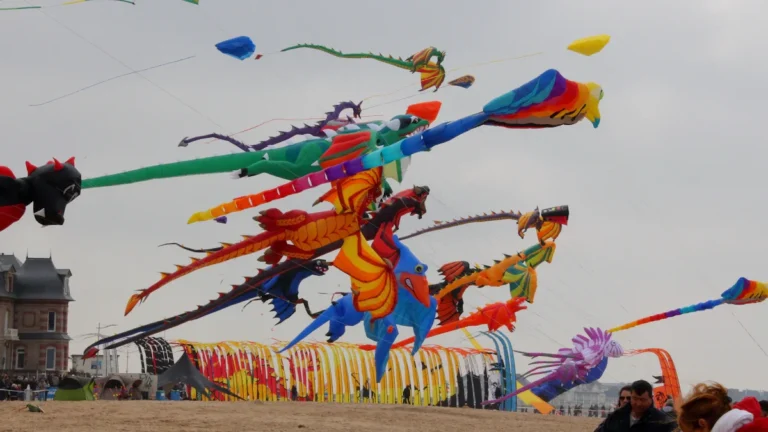 Large, colorful inflatable dragon and creature kites flying over a beach during an international festival.
