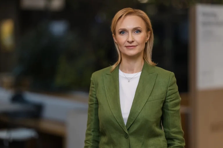 Portrait of a blonde woman wearing a green blazer and white shirt in a blurred office.