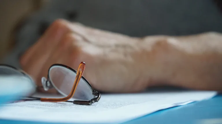 Close-up of reading glasses and an elderly hand resting on a document on a blue table.