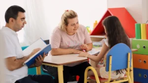 A young child sitting at a desk with two adults who are reviewing documents and providing support in a brightly colored room.