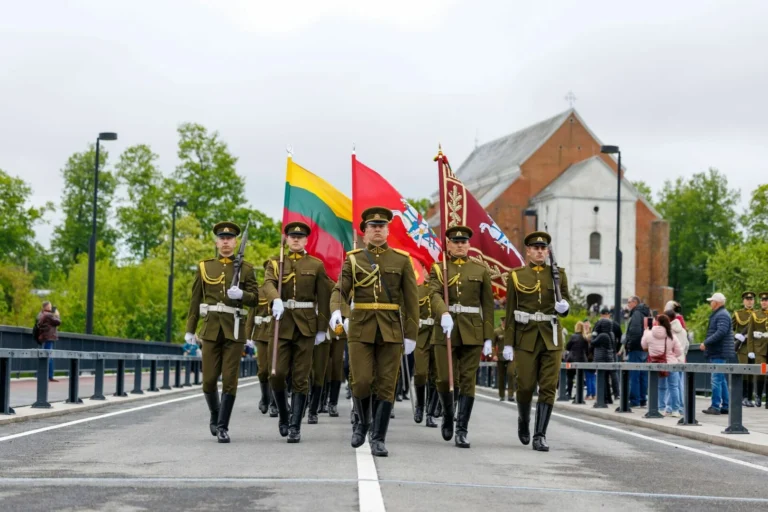 Soldiers in olive uniforms march forward carrying the Lithuanian national flag and military banners.