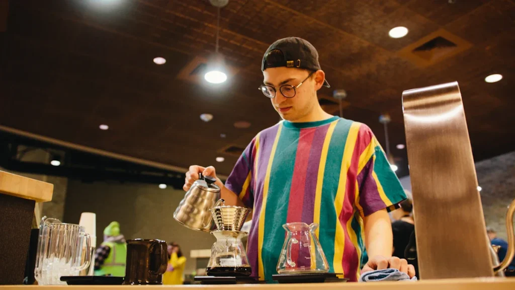 A young man in a colorful striped shirt and glasses carefully prepares a pour-over coffee behind a cafe counter.