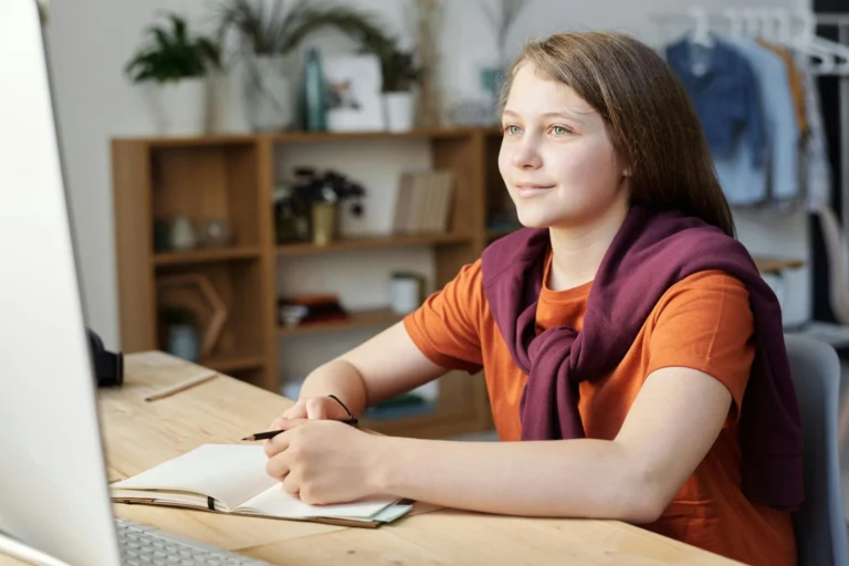 Young girl sitting at a desk looking at a computer monitor with an open notebook.