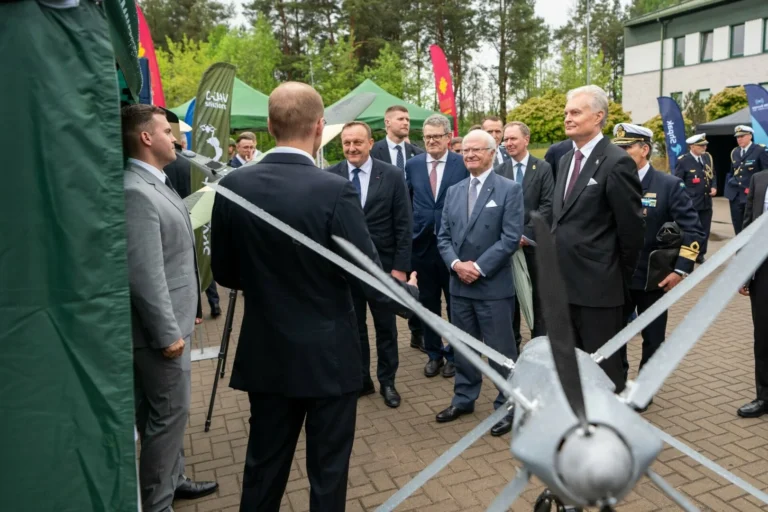 Several men in suits examine a grey unmanned aerial vehicle at an outdoor military exhibition.