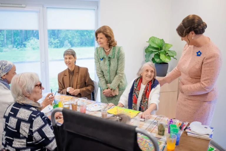 Two women in suits visit elderly people during a painting workshop at a long table.