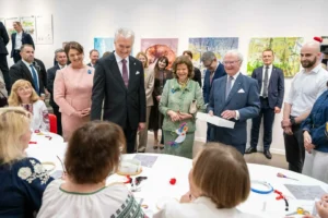 European leaders stand around a table watching women in traditional clothing perform embroidery in a gallery.