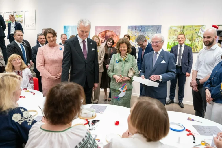 European leaders stand around a table watching women in traditional clothing perform embroidery in a gallery.