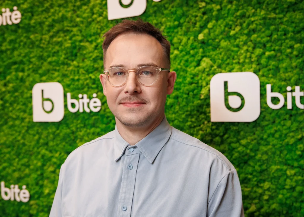 Man with glasses and light blue shirt posing against a green moss wall with logos.