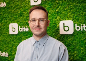 Man with glasses and light blue shirt posing against a green moss wall with logos.