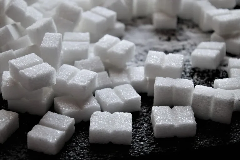 A close-up pile of white granulated sugar cubes scattered on a dark textured surface.