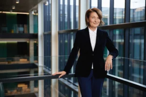 A smiling woman in a navy velvet blazer and white shirt stands in a modern glass office.