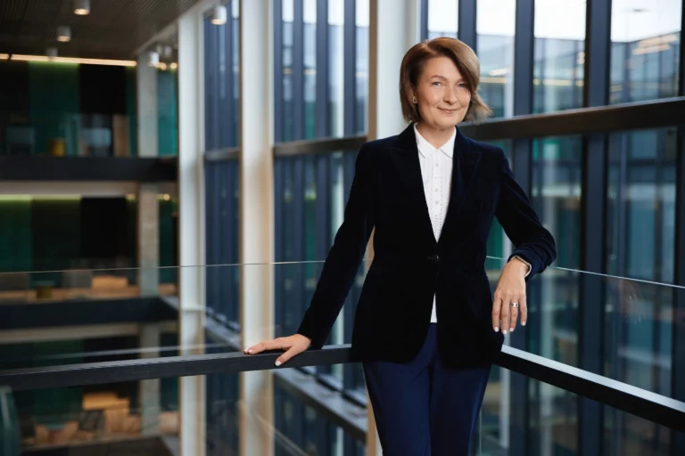 A smiling woman in a navy velvet blazer and white shirt stands in a modern glass office.