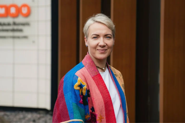 Woman with short blonde hair smiling in a colorful patchwork jacket outside a university building.