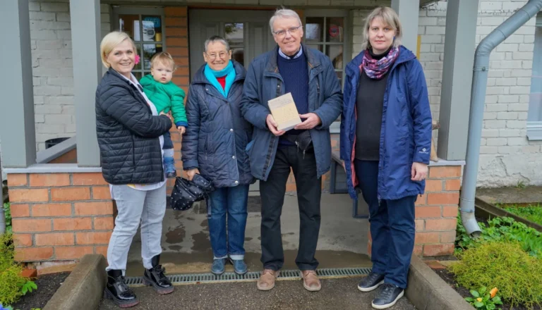 Group of four adults and a toddler posing outside a building while holding a book.