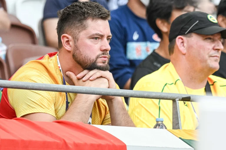 Bearded man in yellow shirt watching intensely with hands under his chin from stadium seating.