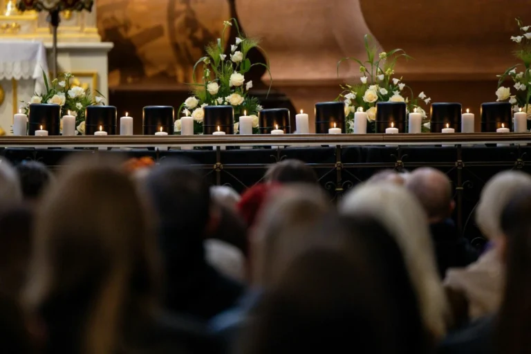 Black funeral urns and lit candles arranged with white roses behind a blurred crowd of people.