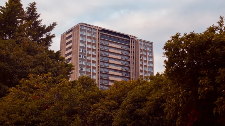 A large white residential apartment building in London partially framed by green trees during the day.