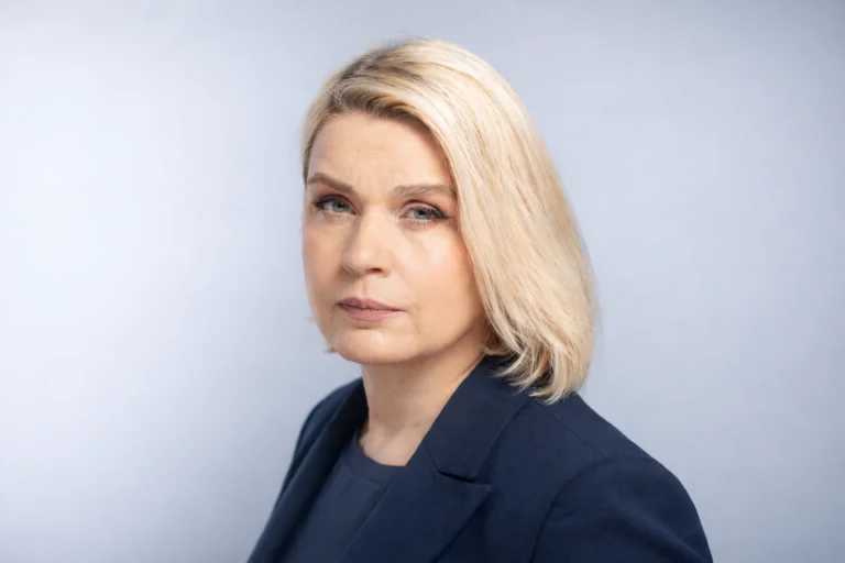 Blonde woman with a serious expression wearing a navy blazer against a light studio background.