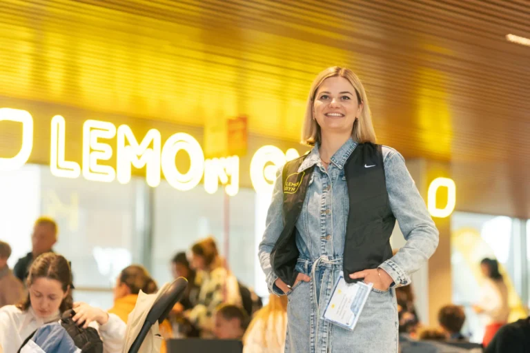 Smiling woman in a denim dress and black vest stands before a neon gym sign.