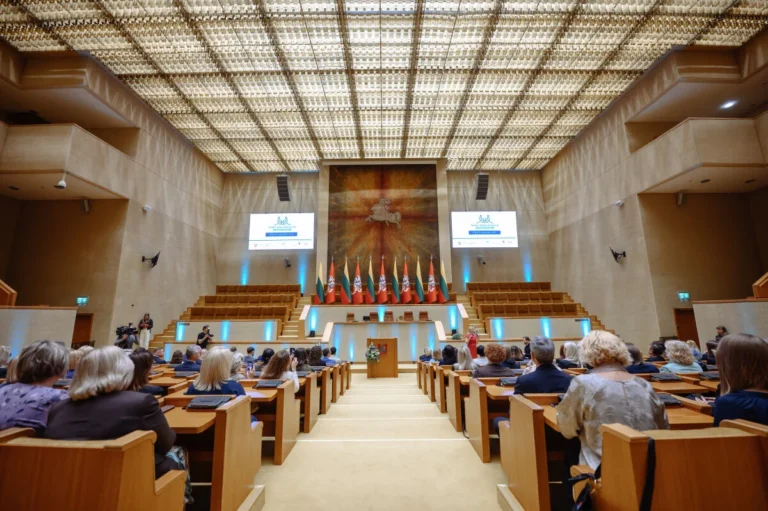 Crowded assembly hall with wooden desks facing a stage featuring several national Lithuanian flags.